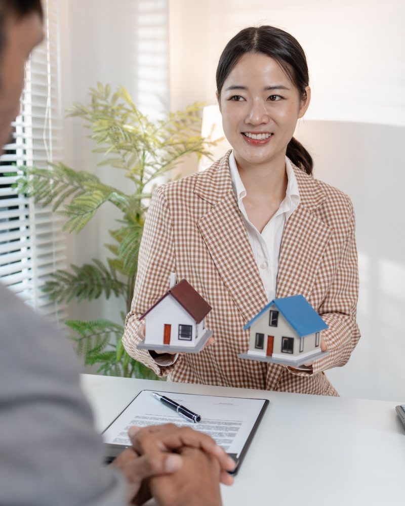 A real estate agent discusses property contracts with a client, using house models and a calculator to explain terms. The scene highlights negotiation, decision-making, and financial planning.